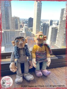 Maurice and Madame Rigolotte, two wooden puppets, in front of a window, with Chicago sky scrappers in the background