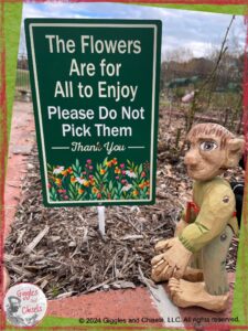 A picture of Pierre Qui Roule, a wooden puppet, in front of sign, saying The flowers are for all to enjoy. Please do not pick them. Thank you.